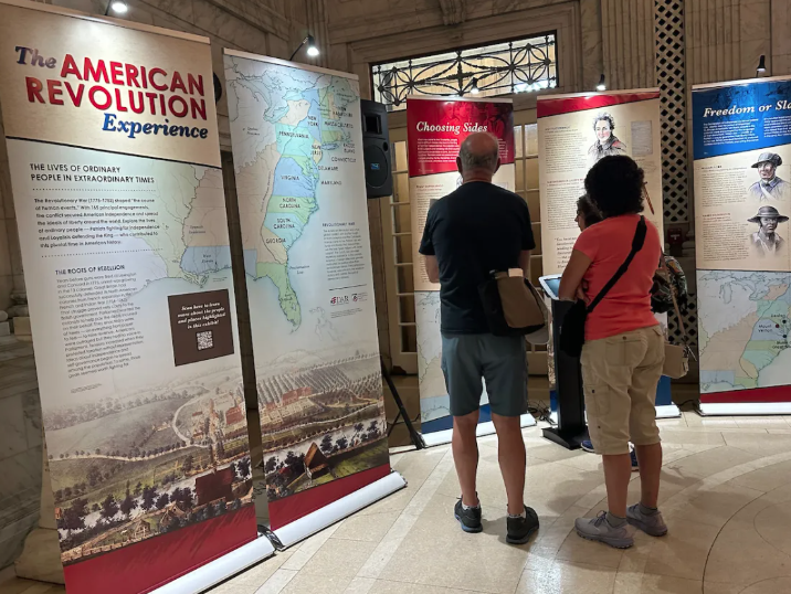 A man in a black shirt and gray shorts and a woman in a pink shirt and tan pants standing and observing banners in an exhibit